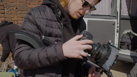 vista de cerca del trabajador de producción pelirrojo con gorra de lana y gafas de sol configurando una cámara en la calle