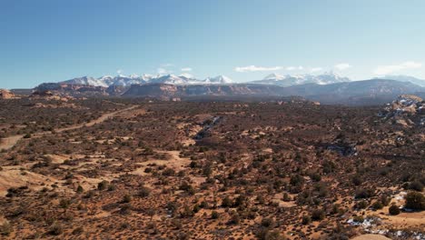 a high-flying drone shot over a remote dirt road cutting through the vast and unique desert land near moab, utah, with the snowy rocky mountains towering in the distance