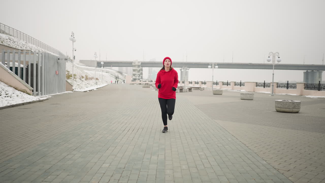 Athlete jogging on interlocked pavement outdoors in winter, surrounded by snow-covered ground, modern urban structures like a bridge, lamp posts, benches, and serene cold-weather scenery