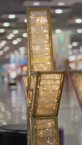 primer plano de intrincados marcos dorados con patrones detallados iluminados por una cálida luz brillante, rodeados de luces blancas parpadeantes, el fondo muestra el interior borroso de un centro comercial con pisos reflectantes