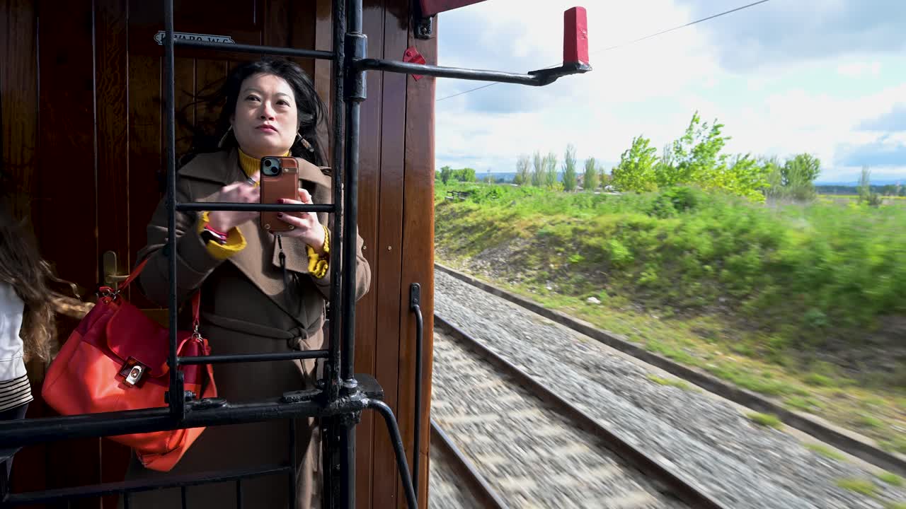 A woman uses her smartphone to take a selfie on a vintage wooden train journey near Madrid, Spain. The trip offers an authentic historical railway experience.