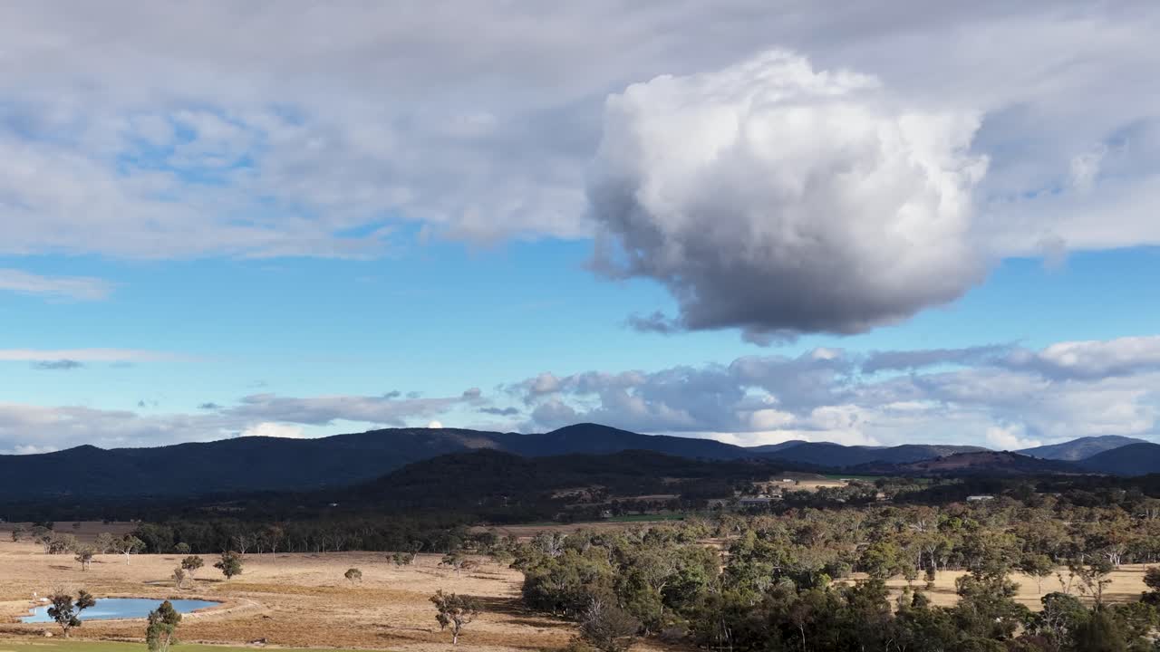 A single large cloud drifts across a sunlit rural landscape with rolling hills, fields, and distant mountains under partly cloudy skies. Static wide shot