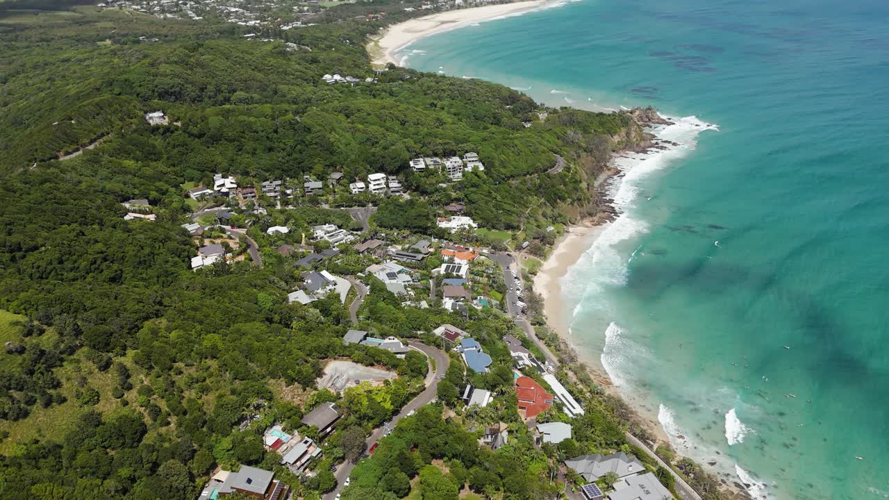 Aerial tilt up of Byron bay with sandy beach and turquoise water. Luxury houses and villas on hillside in Australia. Sunny day with clouds at sky.