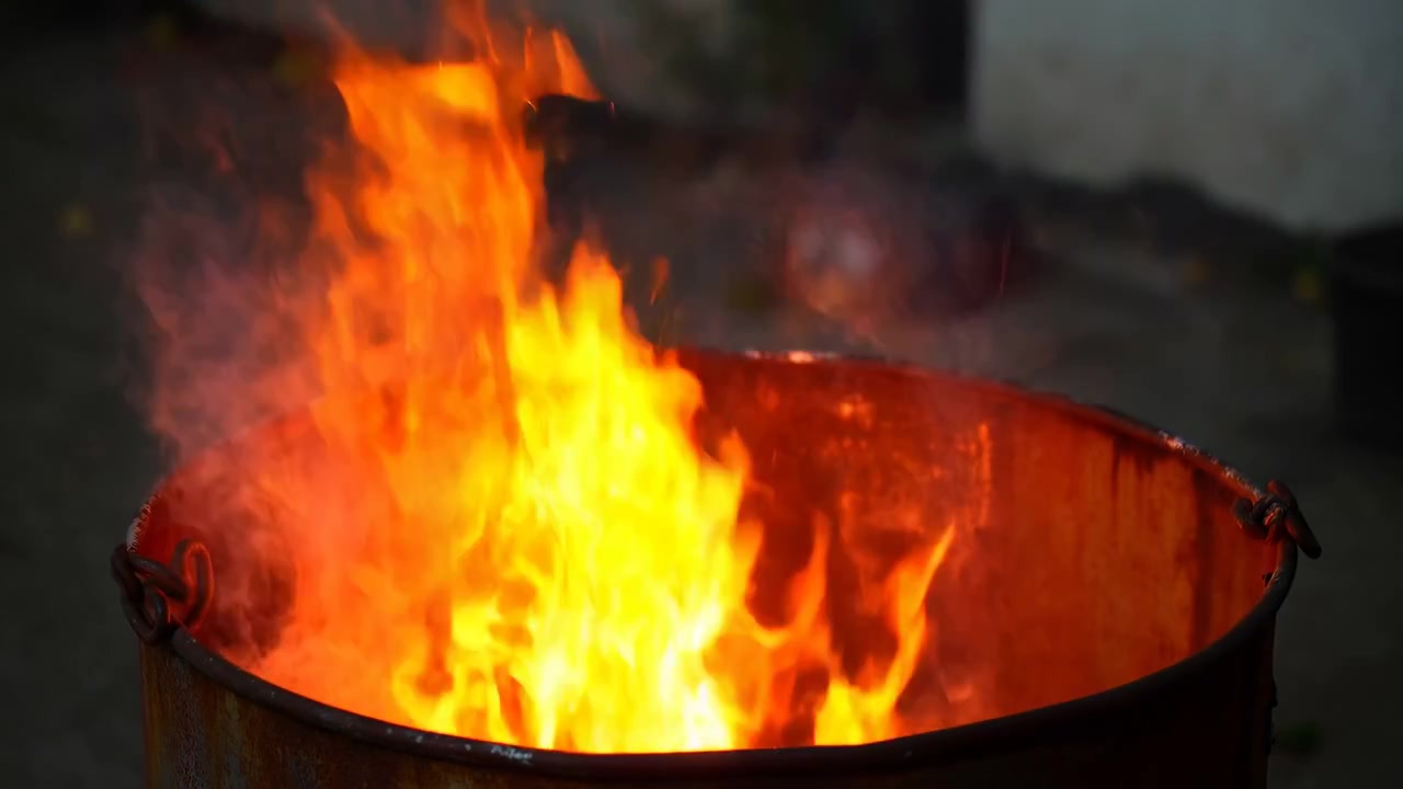 Flammes orange vives dans un baril métallique rouillé, flambant à l'extérieur au crépuscule avec une épaisse fumée et des braises incandescentes.