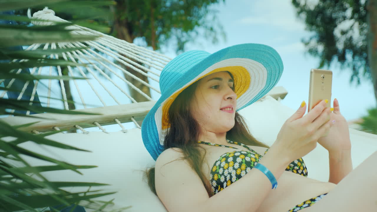 비키니를 입고 챙이 넓은 밝은 모자를 쓴 젊은 검은 머리 소녀가 해먹에서 쉬고 있습니다.