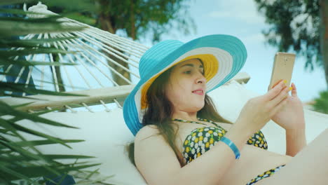 una joven de cabello oscuro en bikini y con un sombrero brillante de ala ancha descansa en una hamaca a