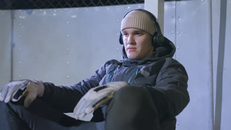 athlete resting against goalpost, shaking head to music through headphones during night training session, wearing gloves and beanie, with illuminated background