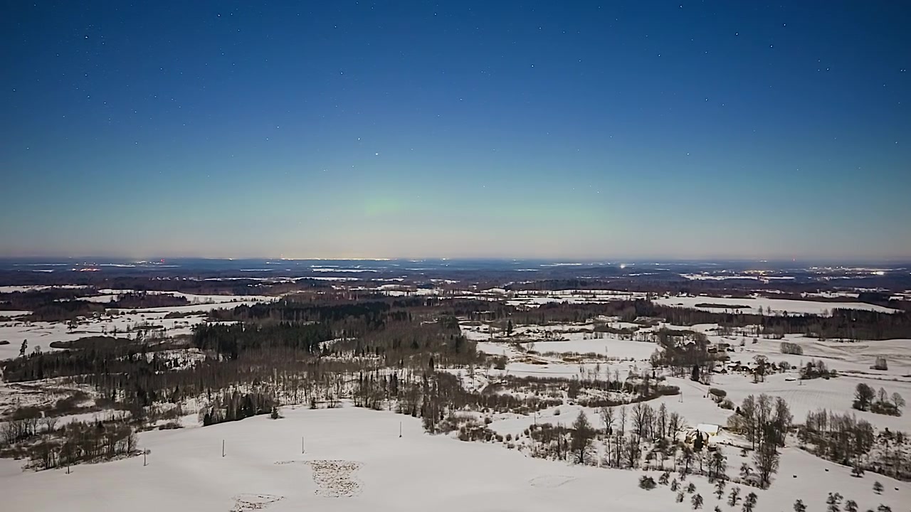 Bright green northern lights illuminating the starry night sky over a snow-covered countryside in aerial time lapse