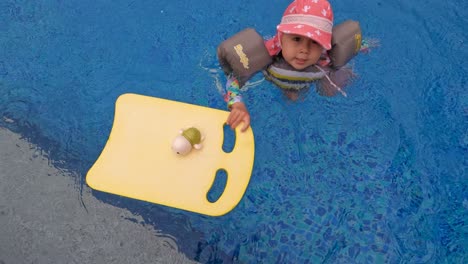 happy little toddler girl swimming in a pool despite of rain playing with toy using foam floating board and jacket for safety