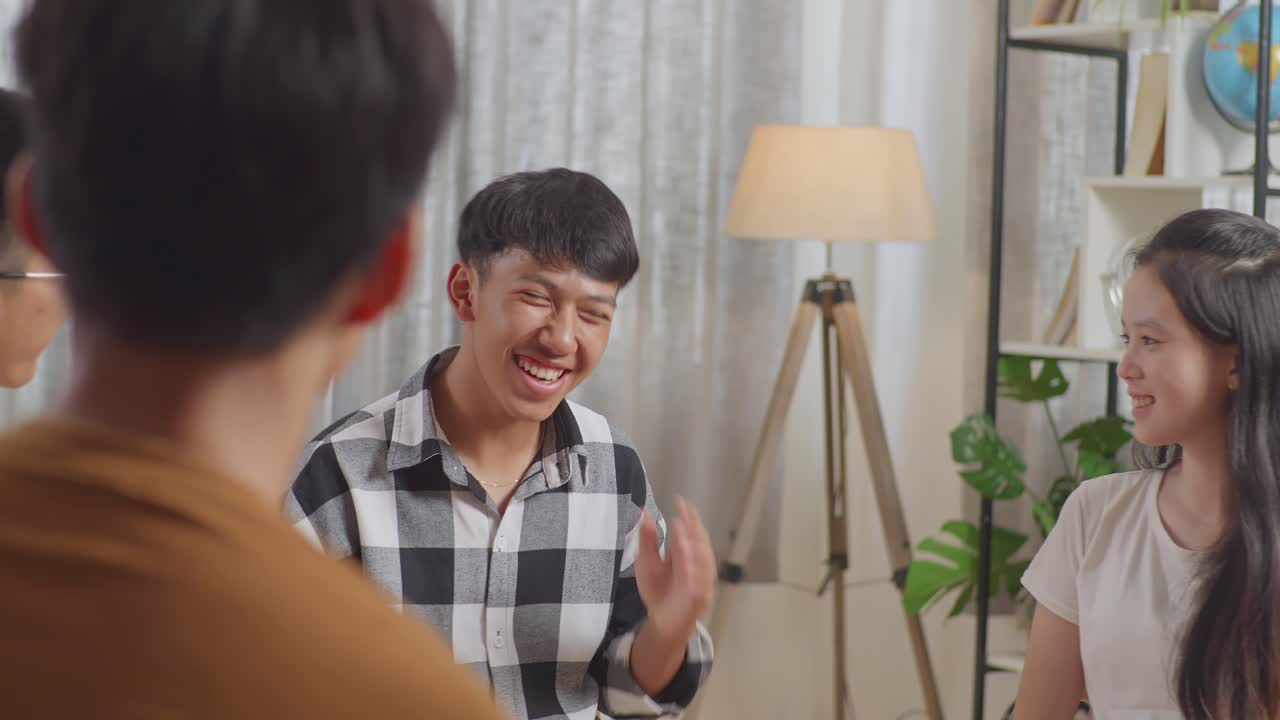 아시안 십대 그룹은 의자에 앉아서 집에서 토론하는 원을 형성합니다. 카레드 셔츠를 입은 소년이 말하고 웃고 있습니다.