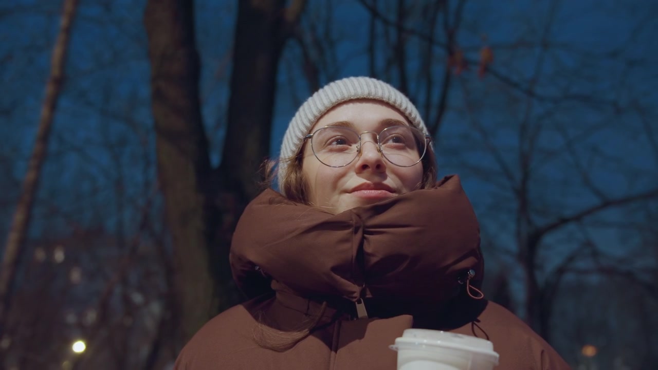 Giovane che sorseggia caffè al parco, indossa un berretto di lana e un piumino spesso, occhiali che riflettono la luce del lampione, tazza fumante e sorriso soddisfatto, alberi notturni e luce calda creano uno stile di vita urbano rilassato.