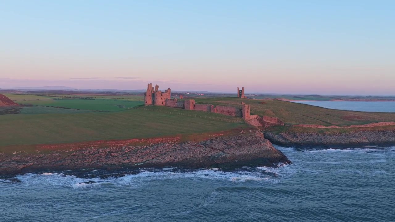 Dramatisk luftfoto, der viser ruinerne af Dunstanburgh Castle på Northumberland-kysten i England under en farverig solopgang.