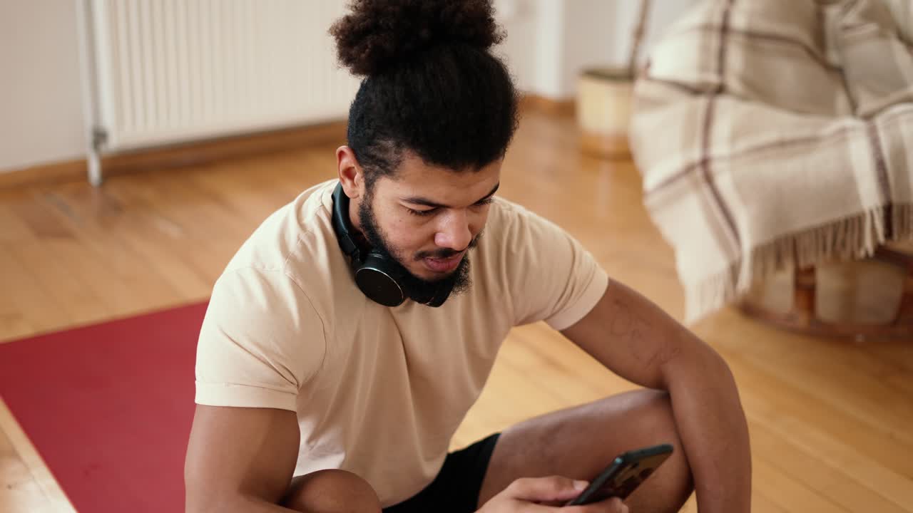 Sportsman sitting on a fitness mat with mobile phone, listening to music with wireless headphones