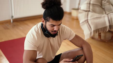 deportista sentado en una alfombra de fitness con teléfono móvil, escuchando música con auriculares inalámbricos