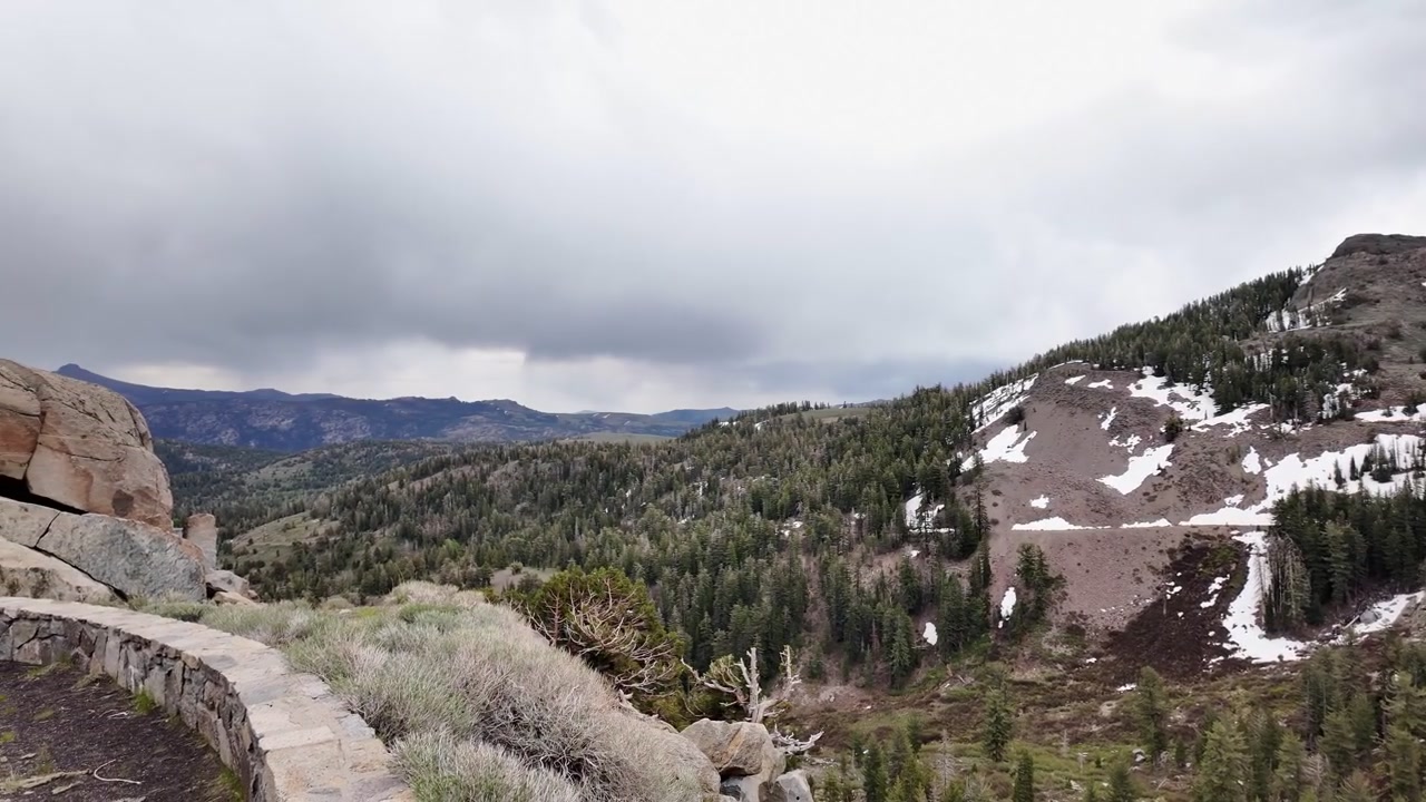 Vista panoramica dal Red Lake Vista Point a Carson Pass in California, che mostra un vasto paesaggio montano con chiazze di neve, foreste di pini e un cielo nuvoloso drammatico
