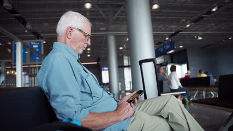 Ein-älterer-Mann-Blickt-Auf-Sein-Telefon,-Während-Er-Am-Flughafen-Geduldig-Auf-Einen-Flug-Wartet