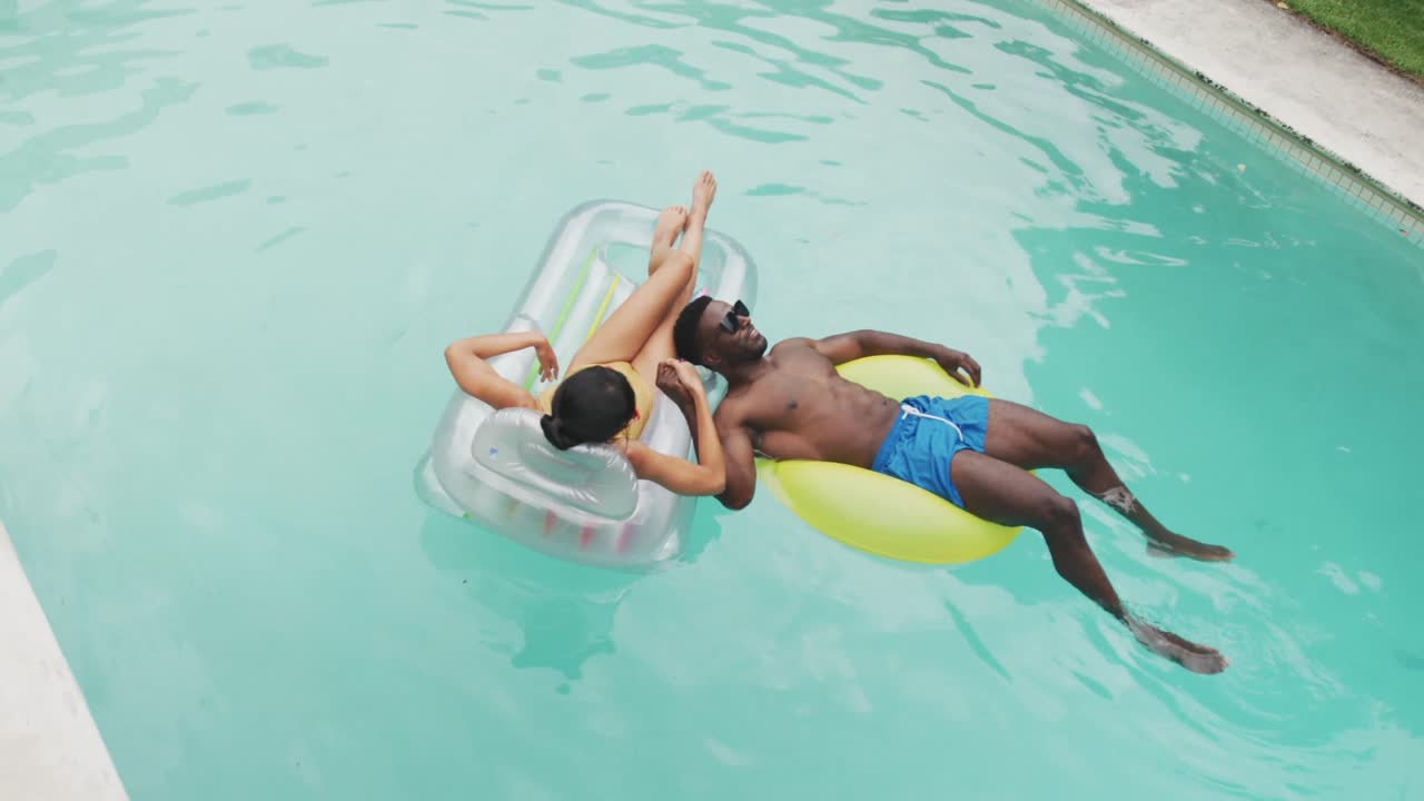 Happy diverse couple wearing swimming suits with inflatables at swimming pool in garden