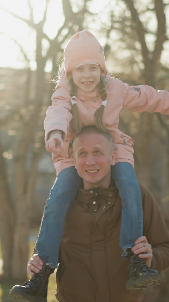 una gioiosa ragazzina con un berretto e una giacca rosa spalanca le braccia mentre si siede sul collo di suo padre mentre camminano attraverso un parco illuminato dal sole. suo padre, indossando una giaccha marrone