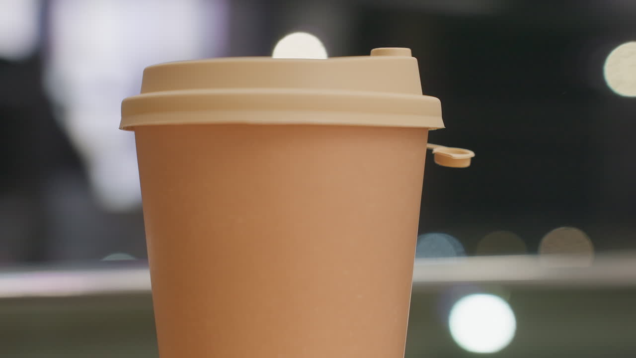 Close up brown takeaway coffee cup with open lid being lifted from wooden table while background shows soft bokeh lights and subtle movements in blurred evening indoor setting