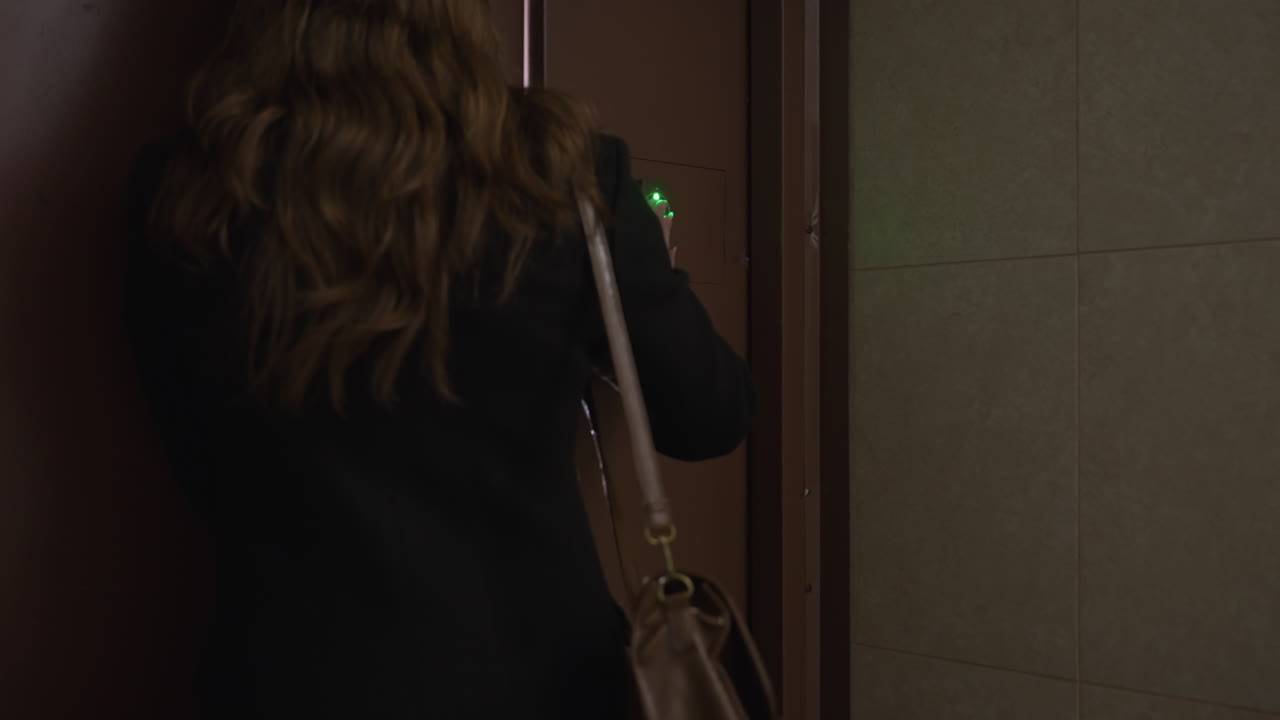 Vista trasera de una mujer con el pelo largo pulsando el acceso inteligente de la puerta, presionando el panel de entrada en la puerta de metal marrón, bolso al hombro, iluminación del pasillo, desenfoque de movimiento, momento de entrada segura al edificio