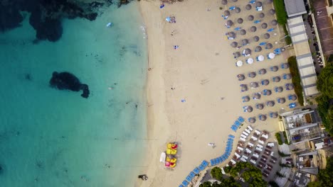 Orbiting,-descending-drone-shot-of-the-Playa-Lletes-beach-resort,-showing-the-beach-umbrellas-lined-in-rows-where-beachgoers-lay-down-for-sunbathing-in-the-Mediterranean-island-of-Mallorca-in-Spain