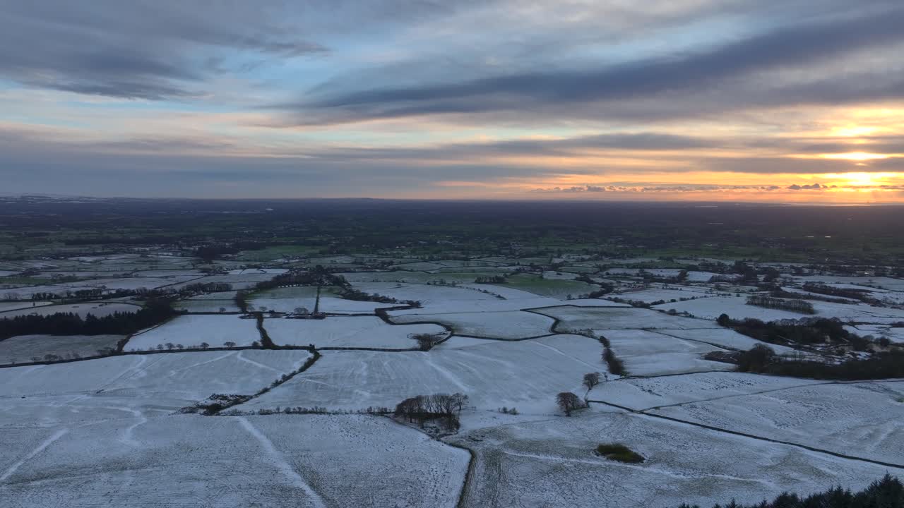 Farmland fields dusted with snow fading out to green. Slow pan revealing setting winter sun. Lancashire, England, UK
