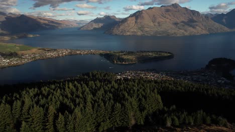 Queenstown,-New-Zealand-aerial-opening-over-tree-line-to-lakeside-town-and-mountain-peak