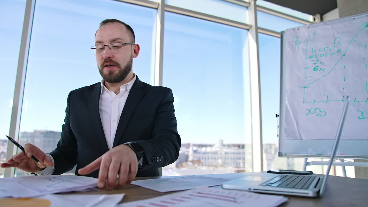 Successful entrepreneur speaking in office. Businessman in glasses and elegant suit sitting at the table in his workplace and talking seriously to someone.