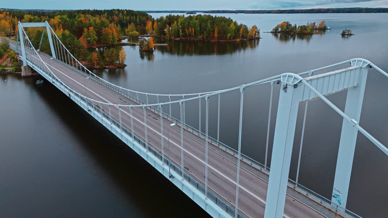 Fixed slow motion high angle shot of a suspension bridge leading from land to archipelago with an autumn forest of red, green, yellow and brown trees