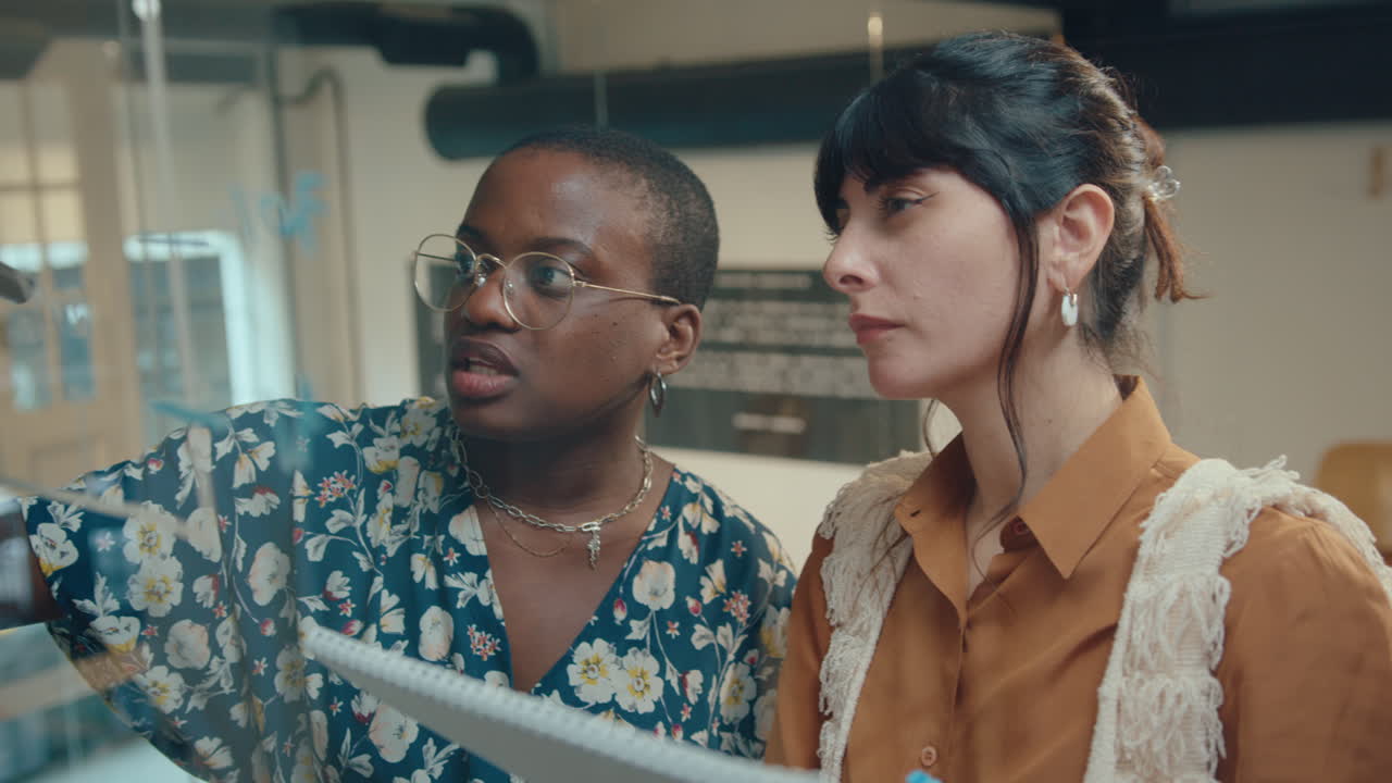 dos mujeres de negocios dibujando gráficos financieros en la pared de una oficina de vidrio