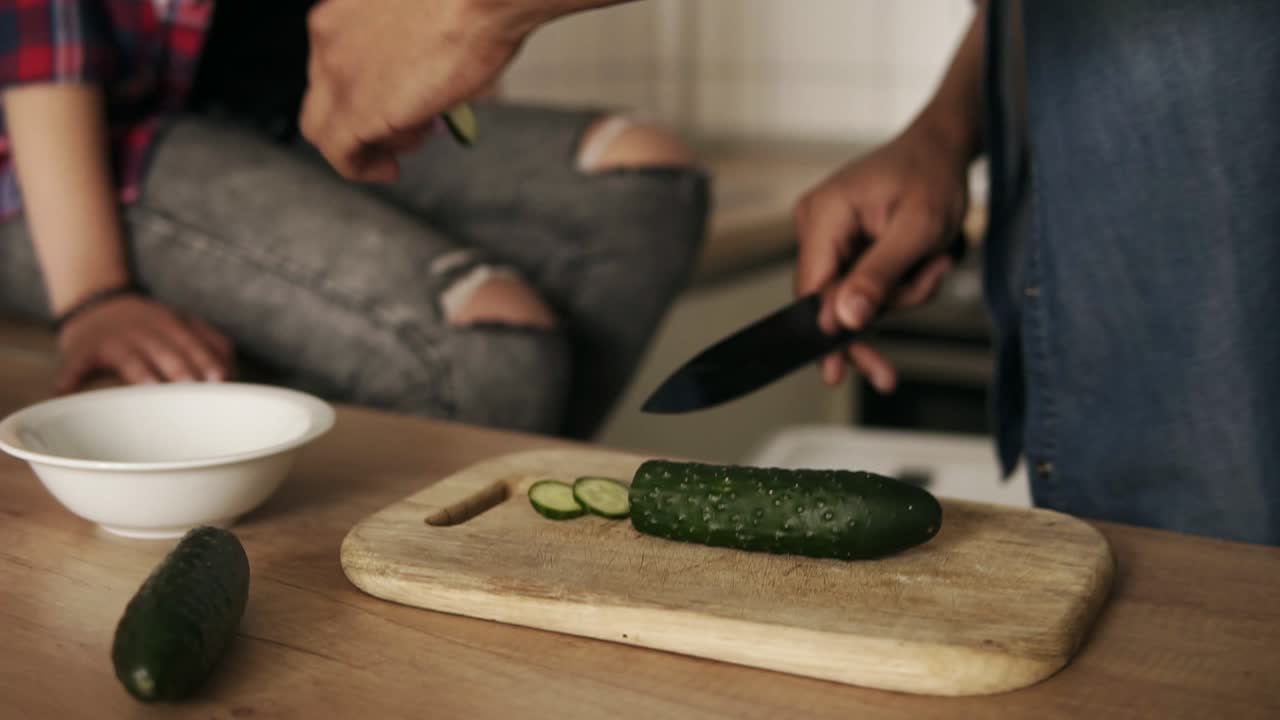 Close up slow motion footage of a young attractive joyful mulatto guy slicing cucumber for salad that he's making for his girlfriend nd himself. Cute couple goals.