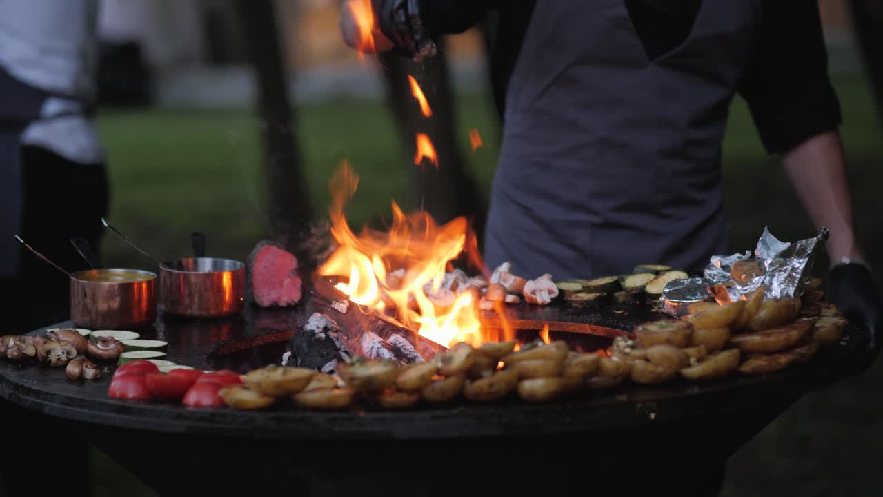 Male chef preparing delicious grilled food on open fire grill in the backyard for the quests during summer evening