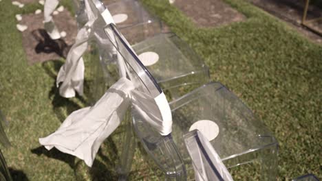 clear chairs decorated with white fabric, placed outdoors on sunlit grass