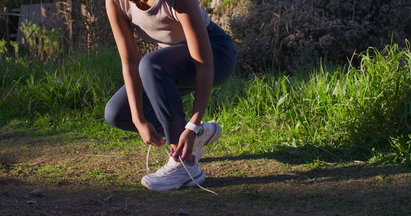 Donna afroamericana che si avvia sul sentiero, preparandosi a correre, allacciandosi i lacci e controllando l'orologio, in abbigliamento sportivo