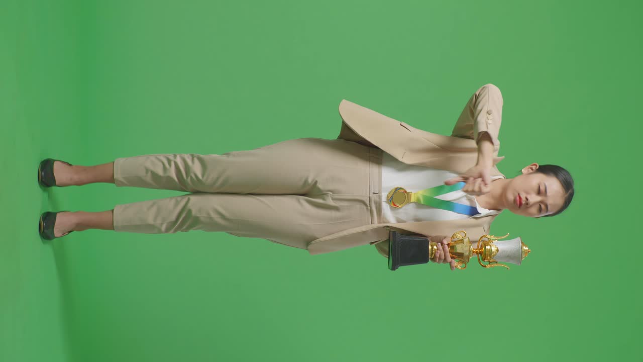 금메달과 트로피를 입은 아시안 사업가 여성의 몸이 가득 차 있습니다. 스튜디오의 녹색 화면 배경에서 패배자를 조롱하기 위해 엄지손가락을 아래로 보여줍니다.