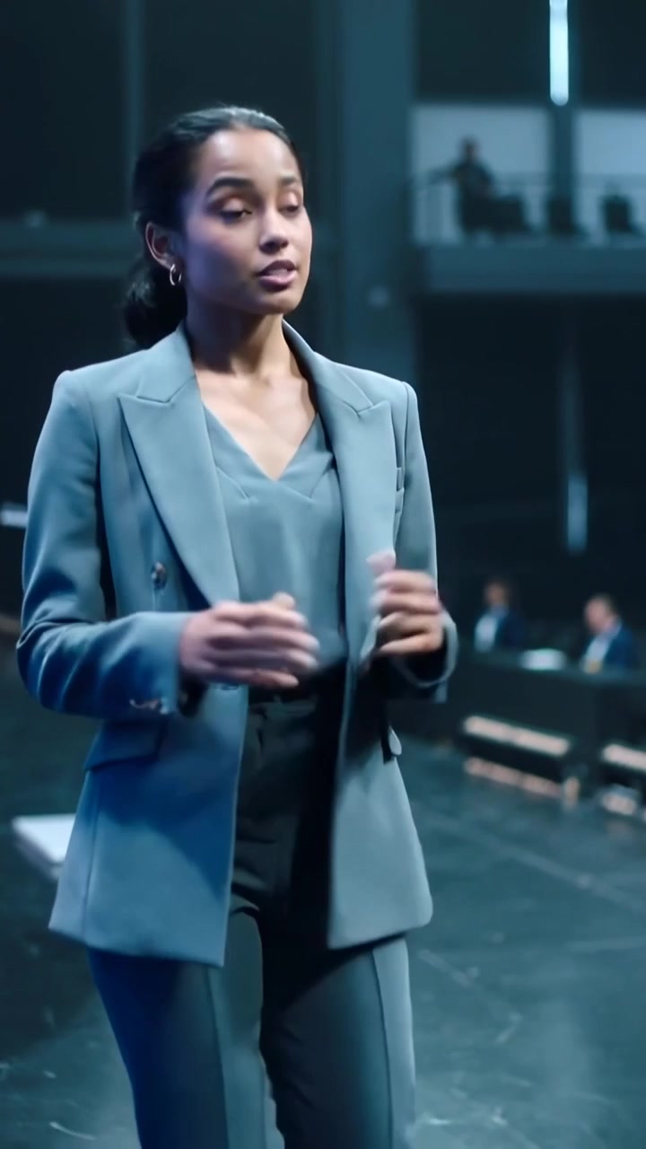 Video verticale: Donna in tailleur color verde acqua che attraversa il palco verso un leggio con due microfoni, rivolgendosi alla folla.