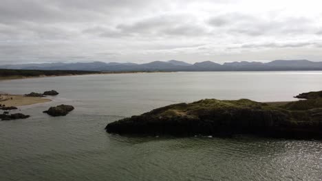 Vista-Aérea-Bajando-A-La-Isla-De-Ynys-Llanddwyn,-Sendero-Costero-De-Anglesey-Con-Montañas-De-Snowdonia-A-Través-Del-Mar-De-Irlanda