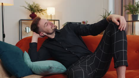 Happy young guy in wireless headphones relaxing lying on sofa at home listening favorite music
