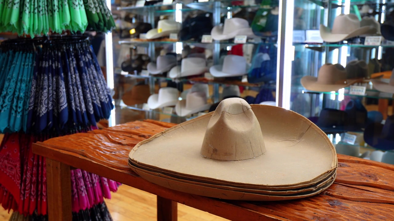 Bases de sombreros apiladas en una mesa de madera en una sombrerería. Formas de fieltro beige sin acabar con pañuelos y sombreros de vaquero al fondo.