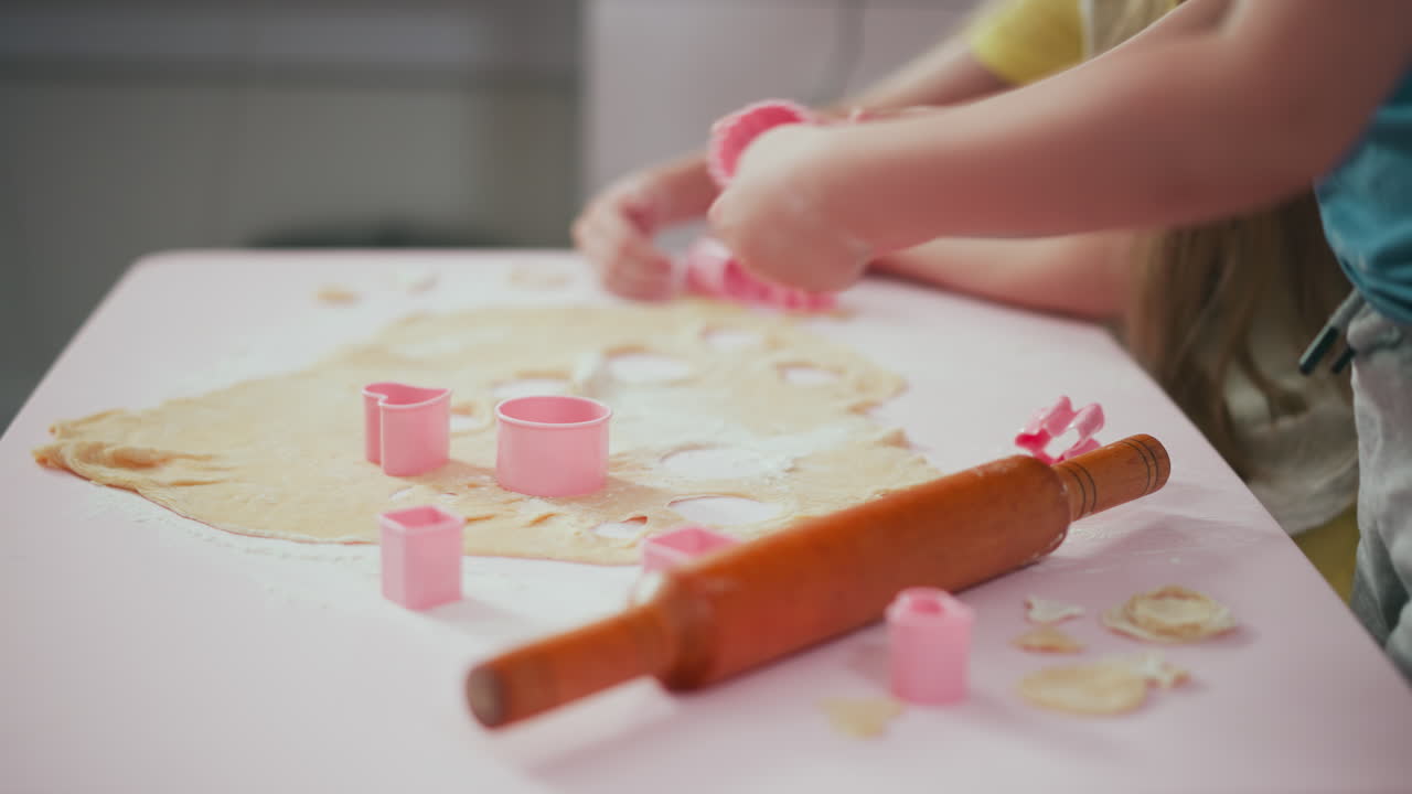 Side view of young children pressing pink cutters into dough on floured table, rolling pin lying nearby, various cutout shapes visible, kids focus on baking