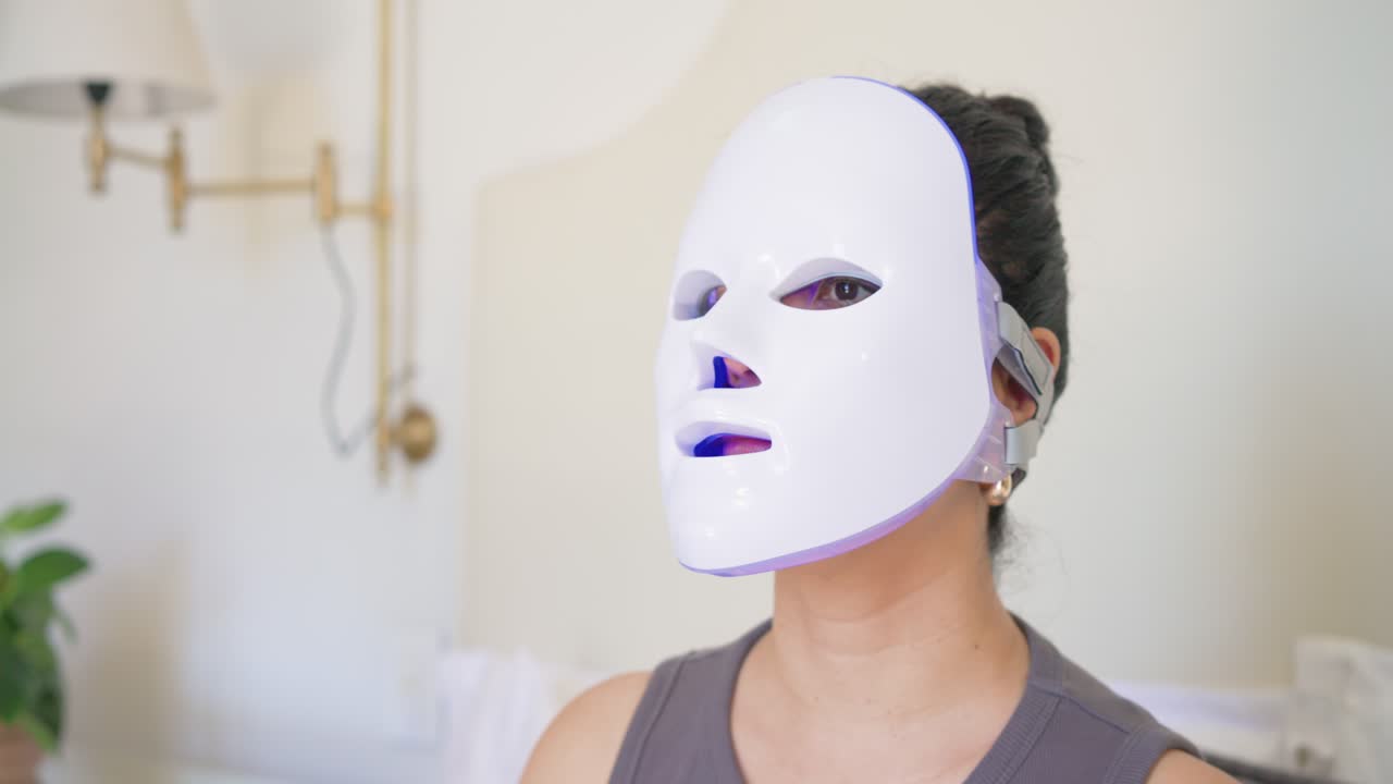 Kvinde løfter hånden, justerer hvid LED-maske i soveværelset med LED'er, der skifter til blå, og sikrer pasformen.