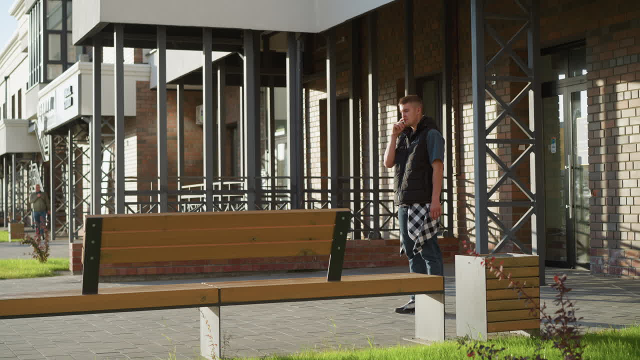 young man standing outside brick building holds cigarette close to lips then pauses and decides to throw it away as distant person walks past in background under sunny daylight in urban setting