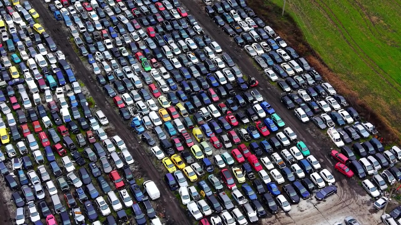 Luftfoto af bilkirkegårdsrækker. Droneoptagelse af hundredvis af parkerede køretøjer i diagonale rækker ved siden af grønne landbrugsarealer i dagslys.