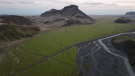 drone shot of a green plain and braided river flowing alongside a rugged mountain in búrfell, iceland, showcasing contrasting landscapes under cloudy skies