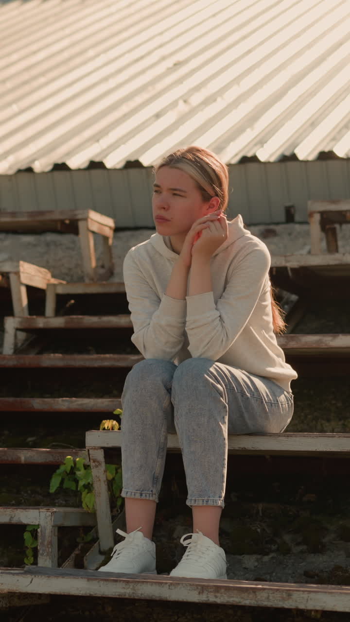 Thoughtful woman in casual attire sits on rustic stadium bleachers with hands joined together, gazing into distance with a pensive and reflective expression