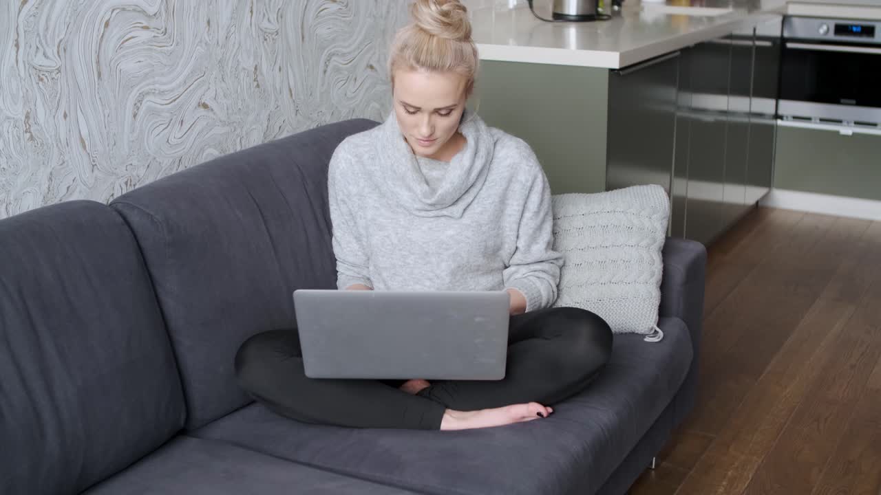 Cheerful young blond woman sitting on couch in living room and using laptop