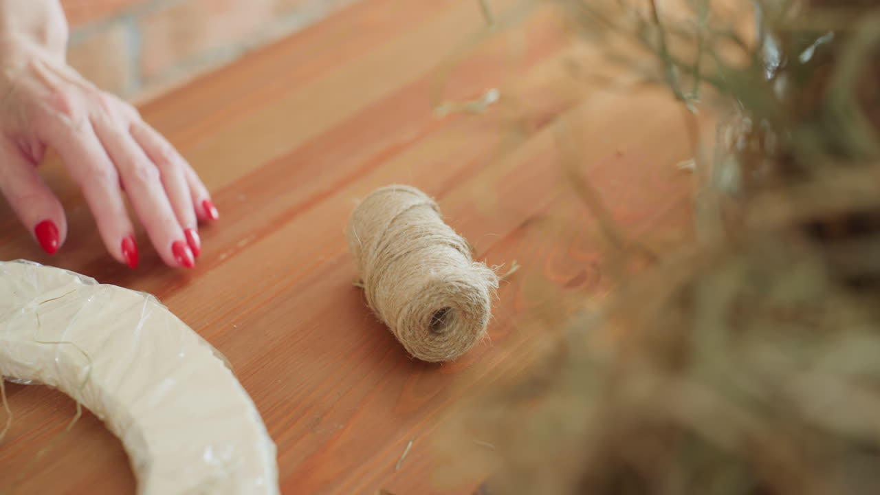 Florist hand with red manicure holds twine roll near wooden box with tools and circular foam wreath base on wooden table in rustic workshop preparing for creative floral arrangement design project