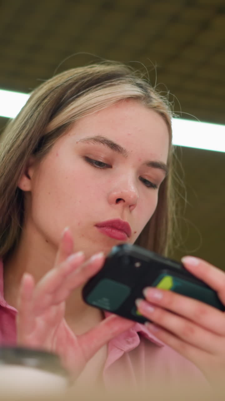 Woman in a pink dress focusing on her phone, seemingly taking a picture of something in front of her, with a bright lights in the background, partial view of a coffee cup is visible