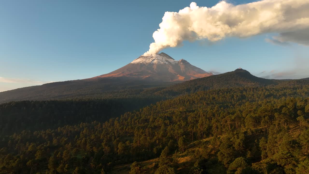 High‑altitude drone push‑in glides above vast dawn forest, revealing Popocatepetl’s snow‑capped cone and thick fumarole against pastel sky—ideal opener for travel, nature or news projects.