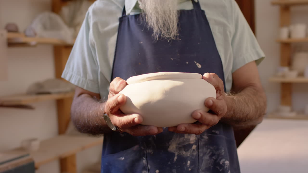 Mid section of biracial potter with long beard holding bowl in pottery studio, slow motion